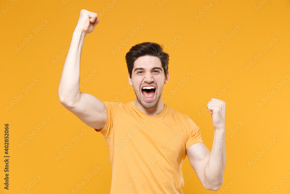 Young smiling unshaved caucasian handsome man 20s in casual basic blank print design t-shirt doing winner gesture with clenching fists saying yes scream isolated on yellow background studio portrait. Young smiling unshaved caucasian handsome man 20s in casual basic blank print design t-shirt doing winner gesture with clenching fists saying yes scream isolated on yellow background studio portrait.