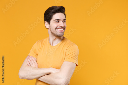 Young smiling unshaved caucasian handsome man 20s years old wearing casual basic blank print design t-shirt look aside holding hands crossed folded isolated on yellow color background studio portrait.