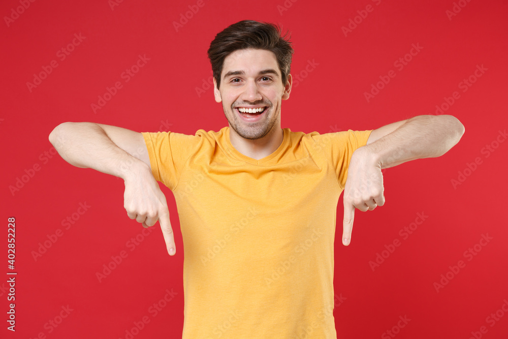 Young smiling unshaved caucasian man 20s in casual blank print design yellow t-shirt point index two fingers down on workspace copy space area look camera isolated on red background studio portrait. Young smiling unshaved caucasian man 20s in casual blank print design yellow t-shirt point index two fingers down on workspace copy space area look camera isolated on red background studio portrait.