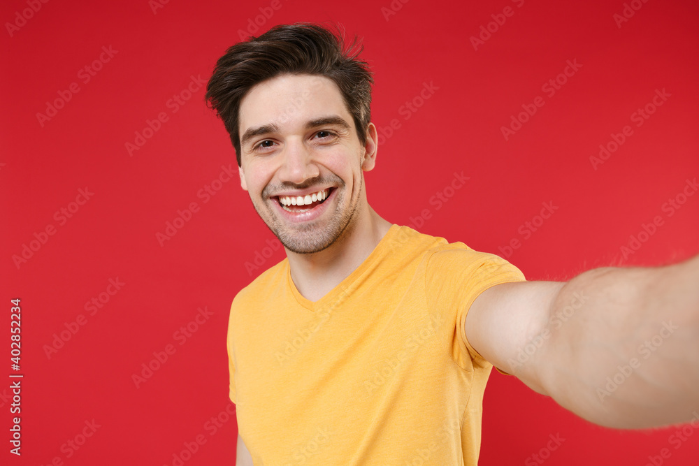 Young smiling fun unshaved friendly caucasian cheerful handsome man 20s wearing casual yellow t-shirt doing close up selfie shot on mobile phone isolated on red color background studio portrait. Young smiling fun unshaved friendly caucasian cheerful handsome man 20s wearing casual yellow t-shirt doing close up selfie shot on mobile phone isolated on red color background studio portrait.