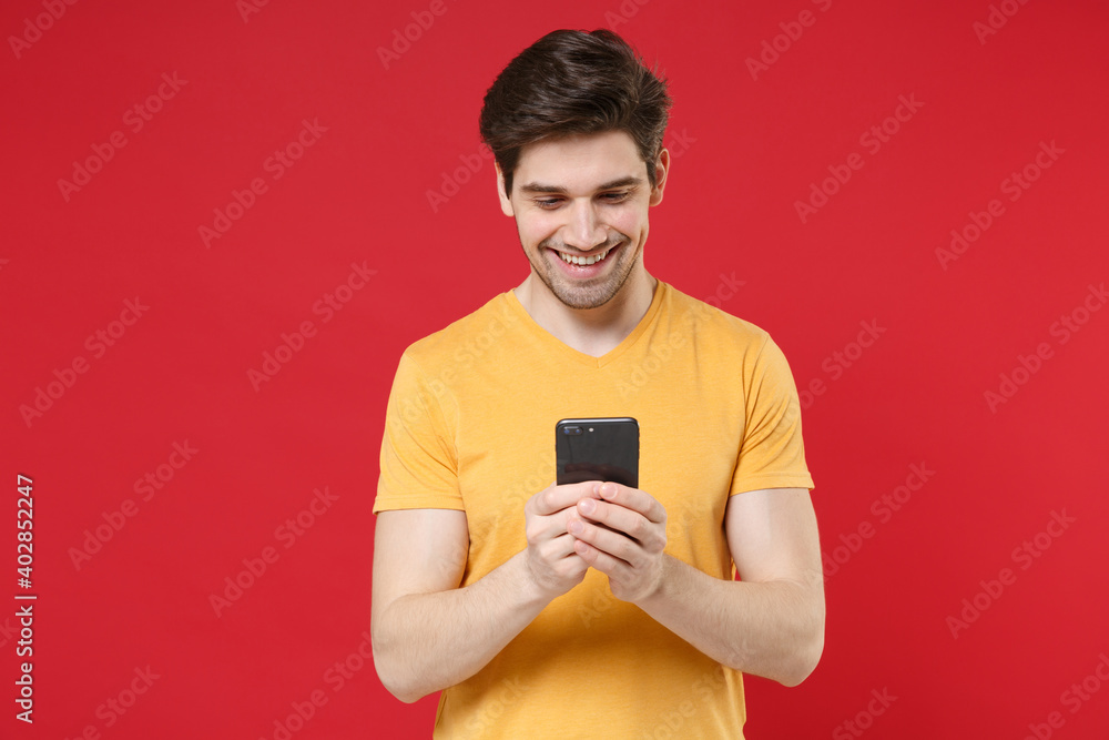 Young smiling caucasian man 20s wearing casual basic yellow t-shirt holding modern mobile cell phone typing chatting surfing internet reading news isolated on red color background studio portrait Young smiling caucasian man 20s wearing casual basic yellow t-shirt holding modern mobile cell phone typing chatting surfing internet reading news isolated on red color background studio portrait