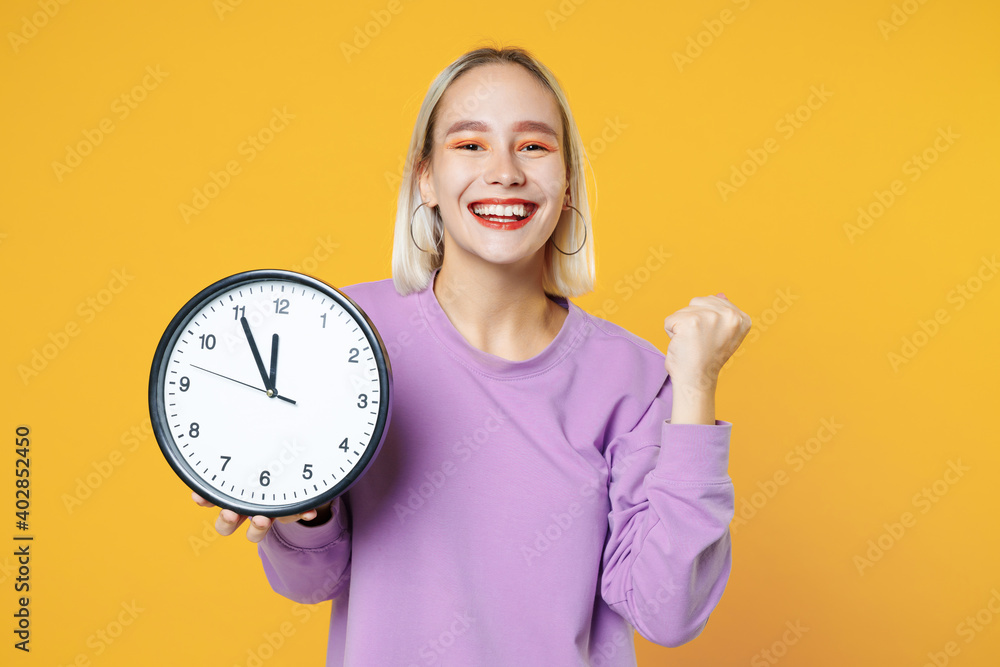 Young blonde caucasian escited woman 20s bob haircut makeup wearing casual basic purple shirt holding in hands clock doing winner gesture cleching fist isolated on yellow background studio portrait. Young blonde caucasian escited woman 20s bob haircut makeup wearing casual basic purple shirt holding in hands clock doing winner gesture cleching fist isolated on yellow background studio portrait.