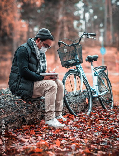 Man with protectiv mask sitting outside on a stock writing in a book, with his bike next to him.  photo