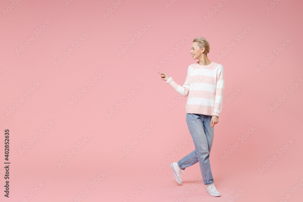 Full length young fashionable blonde woman 20s with short haircut in casual knitted sweater jeans standing pointing index finger aside looking back isolated on pastel pink background studio portrait. Full length young fashionable blonde woman 20s with short haircut in casual knitted sweater jeans standing pointing index finger aside looking back isolated on pastel pink background studio portrait.