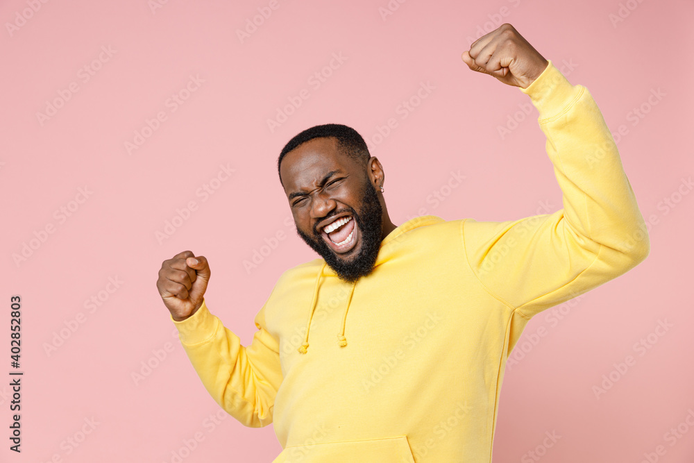 Overjoyed young african american man 20s wearing casual basic yellow streetwear hoodie standing doing winner gesture clenching fists isolated on pastel pink color wall background studio portrait. Overjoyed young african american man 20s wearing casual basic yellow streetwear hoodie standing doing winner gesture clenching fists isolated on pastel pink color wall background studio portrait.