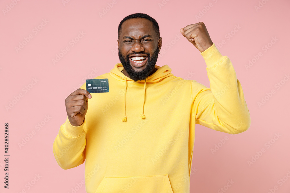 Joyful young african american man 20s in casual yellow streetwear hoodie standing hold credit bank card doing winner gesture looking camera isolated on pastel pink color background studio portrait. Joyful young african american man 20s in casual yellow streetwear hoodie standing hold credit bank card doing winner gesture looking camera isolated on pastel pink color background studio portrait.