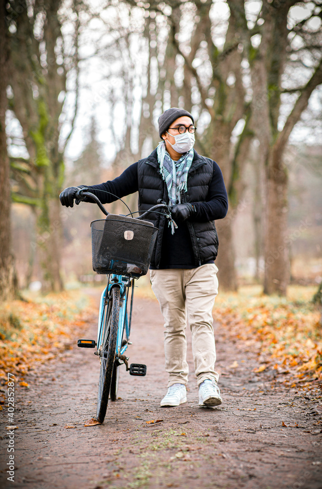 Asian man with protective mask is outside walking with his blue bike. 