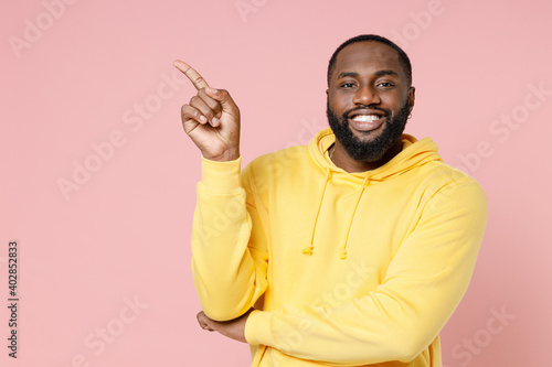 Smiling young african american man 20s in casual basic yellow streetwear hoodie standing pointing index finger aside up on mock up copy space isolated on pastel pink color background studio portrait.