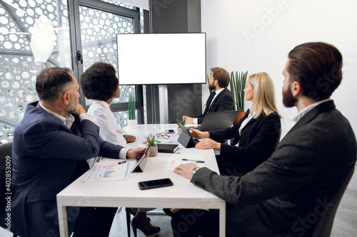 Diverse group of successful multiracial focused businesspeople in the conference room, looking at digital wall screen with white empty copy space. Business meeting concept