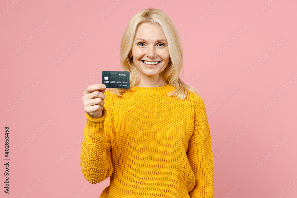 Smiling elderly gray-haired blonde woman lady 40s 50s years old wearing yellow casual sweater standing hold credit bank card looking camera isolated on pastel pink color background studio portrait. Smiling elderly gray-haired blonde woman lady 40s 50s years old wearing yellow casual sweater standing hold credit bank card looking camera isolated on pastel pink color background studio portrait.