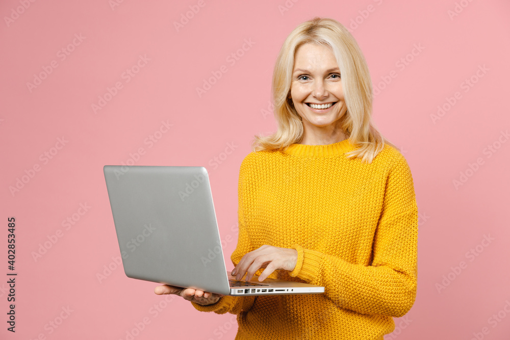 Smiling cheerful elderly gray-haired blonde woman lady 40s 50s years old wearing yellow casual sweater standing working on laptop pc computer isolated on pastel pink color background studio portrait. Smiling cheerful elderly gray-haired blonde woman lady 40s 50s years old wearing yellow casual sweater standing working on laptop pc computer isolated on pastel pink color background studio portrait.