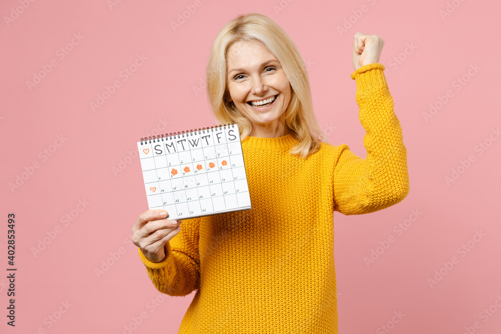 Happy elderly gray-haired blonde woman 40s in sweater doing winner gesture hold female period calendar checking menstruation days isolated on pink background. Medical healthcare gynecological concept. Happy elderly gray-haired blonde woman 40s in sweater doing winner gesture hold female period calendar checking menstruation days isolated on pink background. Medical healthcare gynecological concept.