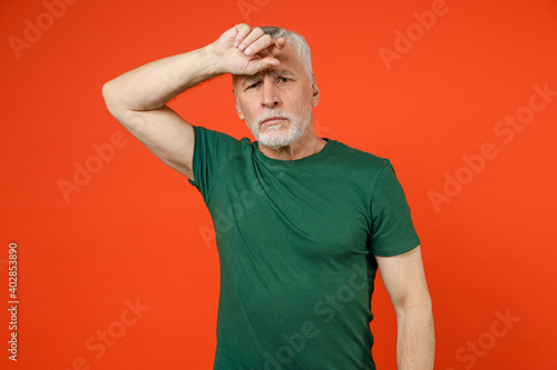 Upset exhausted tired elderly gray-haired mustache bearded man wearing basic green t-shirt standing put hand on head looking camera isolated on bright orange color wall background studio portrait.