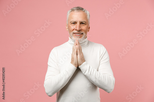 Smiling pleading elderly gray-haired mustache bearded man wearing casual basic white turtleneck standing hold hands folded in prayer looking camera isolated on pastel pink background studio portrait. © ViDi Studio