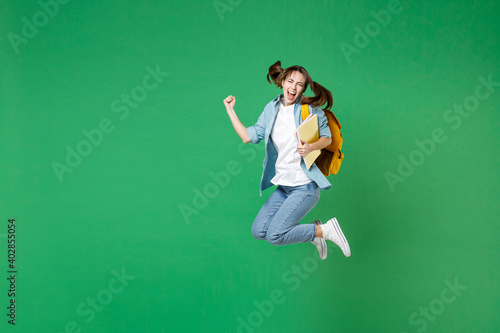 Full length happy young woman student in shirt backpack hold notebooks jumping doing winner gesture isolated on green background studio portrait. Education in high school university college concept.