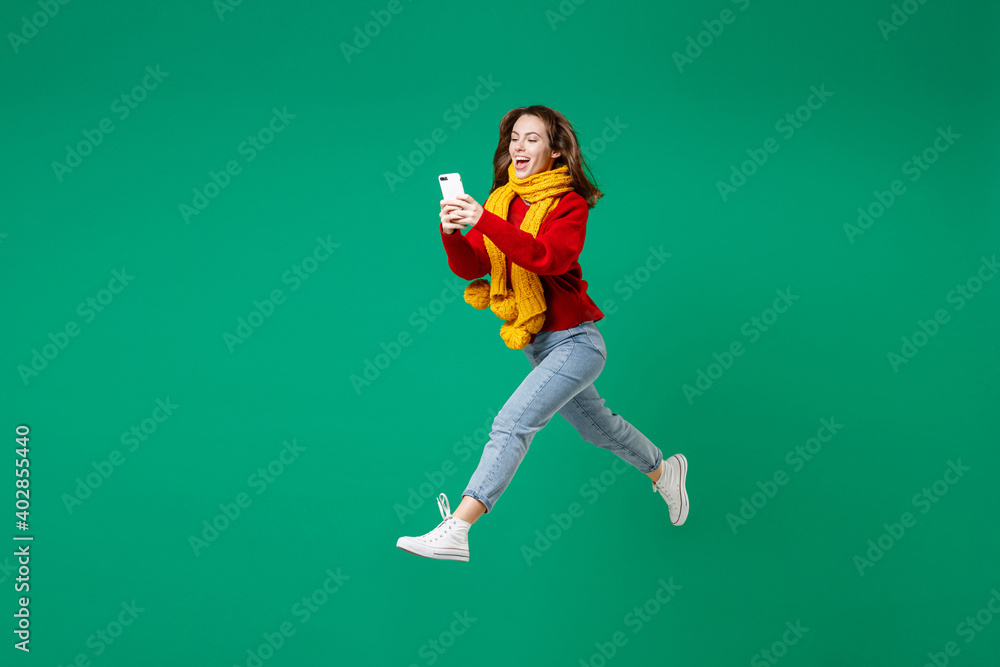 Full length side view of cheerful funny young woman 20s wearing knitted red sweater yellow scarf jumping using mobile cell phone typing sms message isolated on green color background studio portrait. Full length side view of cheerful funny young woman 20s wearing knitted red sweater yellow scarf jumping using mobile cell phone typing sms message isolated on green color background studio portrait.