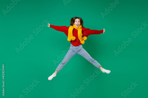 Full length of excited laughing young brunette woman 20s in casual knitted red sweater yellow scarf jumping spreading hands and legs looking camera isolated on green color background studio portrait.