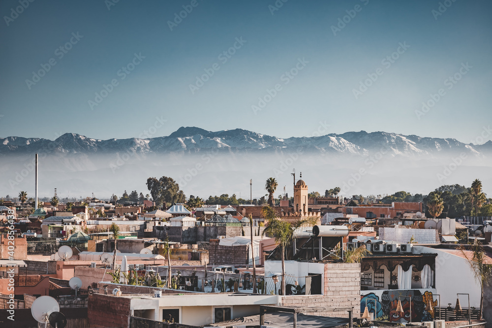 Panoramic sunset view of Marrakesh Medina and the snow capped Atlas mountains in Morocco. View of rooftops of Marrakech close to the city centre. Koutoubia Mosque minaret during Sunrise on blue sky.