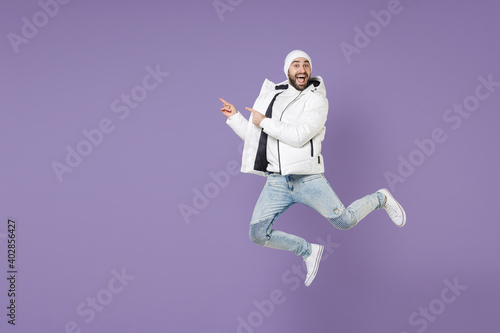 Full length of excited man in warm white padded windbreaker jacket hat jumping pointing index fingers aside isolated on purple background studio portrait. People lifestyle cold winter season concept.