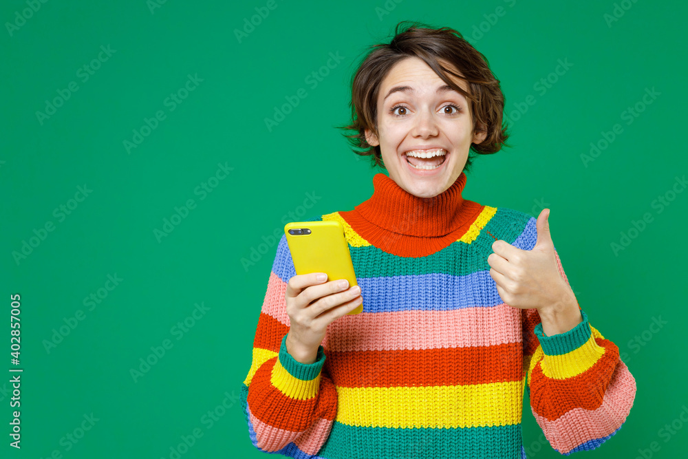 Excited young brunette woman 20s wearing basic casual colorful sweater standing using mobile cell phone typing sms message showing thumb up isolated on bright green color background studio portrait. Excited young brunette woman 20s wearing basic casual colorful sweater standing using mobile cell phone typing sms message showing thumb up isolated on bright green color background studio portrait.