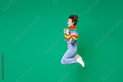 Full length side view of excited young brunette woman 20s years old in basic casual colorful sweater jumping doing winner gesture say yes isolated on bright green color background studio portrait.