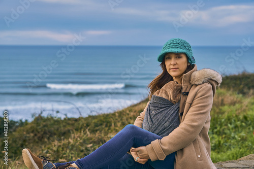 portrait of woman with hat and coat looking at the seascape and cliffs in winter on a cloudy day carrying her newborn baby. concept go out in winter