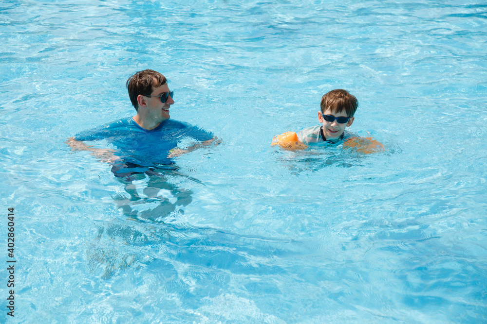Boy and father in Sunglasses with inflatable handcuffs swimming in a pool with blue water. Dad is learning his son to swim and playing in swimming pool. Active healthy lifestyle, water sport activity
