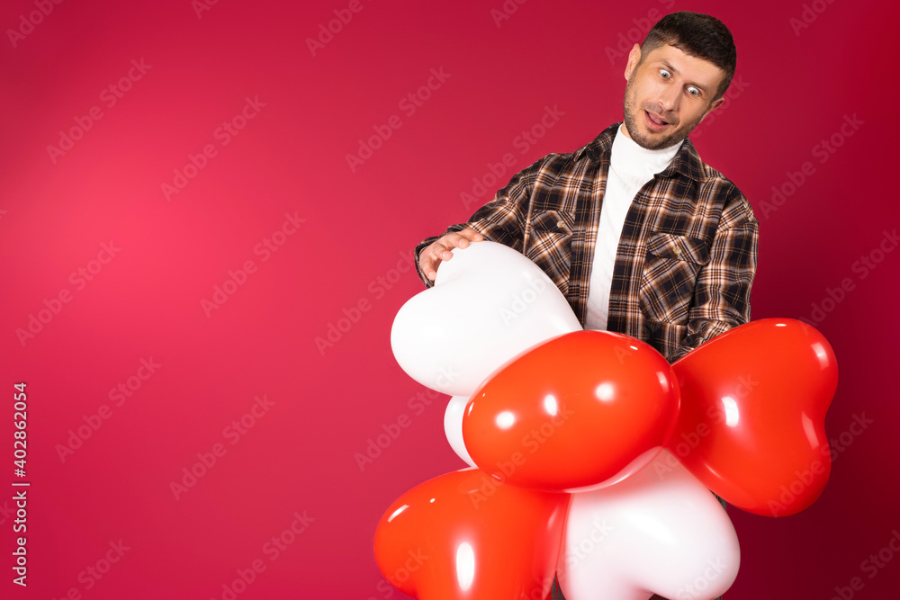 A guy makes faces while holding a bunch of heart-shaped red balloons. Red background and empty sidewall space.