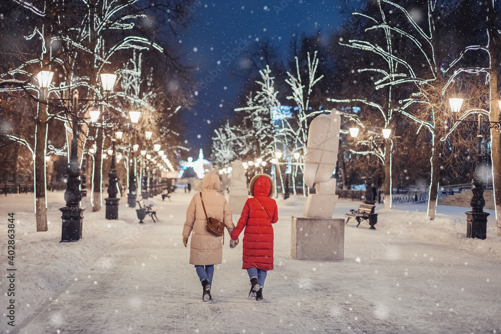 Happy female friends walk in the evening winter park on the sidewalk against the background of Christmas and New Year decorations in Ufa, Russia Happy female friends walk in the evening winter park on the sidewalk against the background of Christmas and New Year decorations in Ufa, Russia