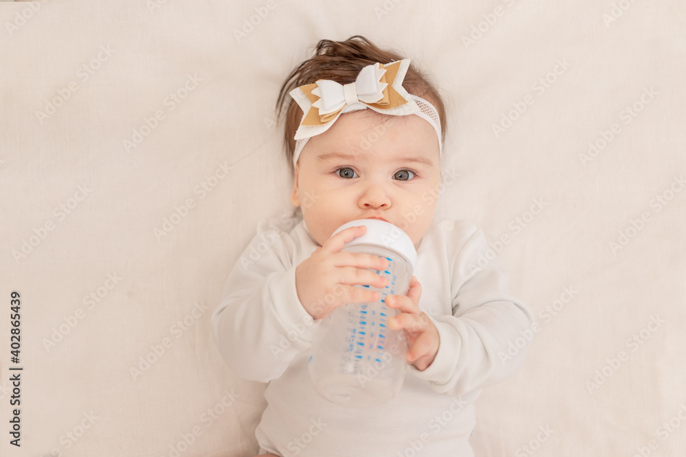 baby six months lies in a crib with a bottle and drinks water in a white bodysuit