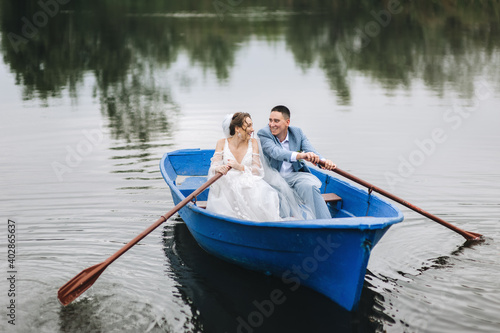 Stylish groom in a suit and a cute bride in a white lace dress are sitting in a wooden boat, walking and swimming on the lake, enjoying the beautiful nature.