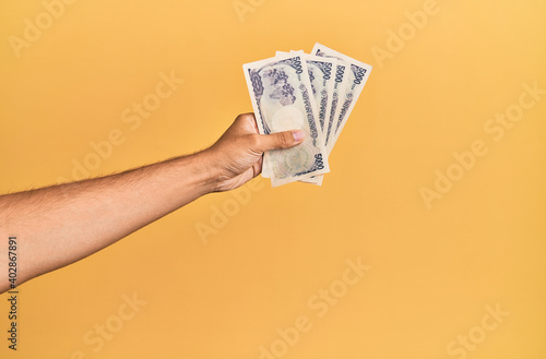 Hand of hispanic man holding japanese yen banknotes over isolated yellow background.