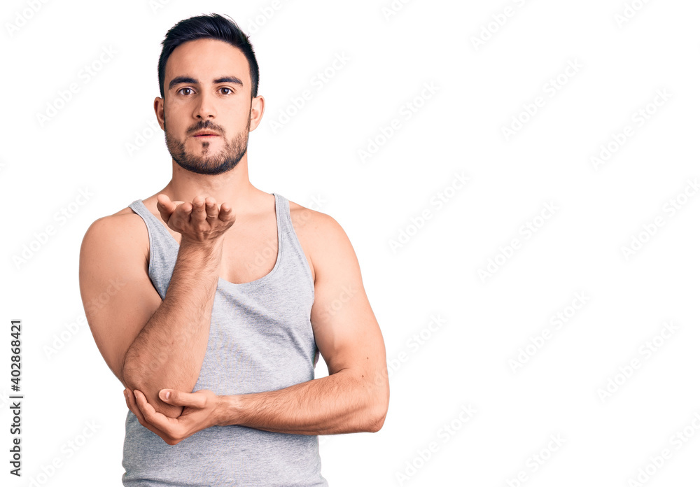 Young handsome man wearing swimwear and sleeveless t-shirt looking at the camera blowing a kiss with hand on air being lovely and sexy. love expression. Young handsome man wearing swimwear and sleeveless t-shirt looking at the camera blowing a kiss with hand on air being lovely and sexy. love expression.