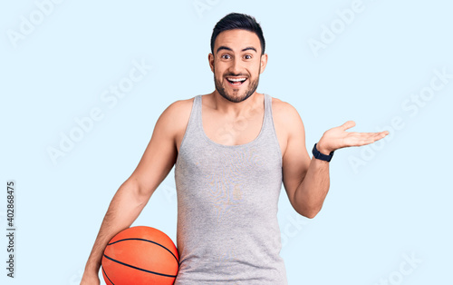 Young handsome man holding basketball ball celebrating victory with happy smile and winner expression with raised hands