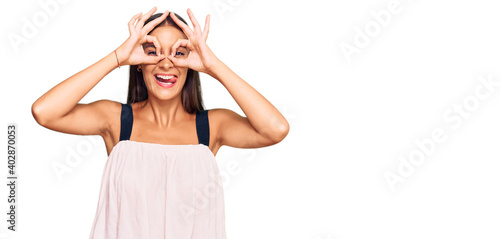 Young hispanic woman wearing casual clothes doing ok gesture like binoculars sticking tongue out, eyes looking through fingers. crazy expression.