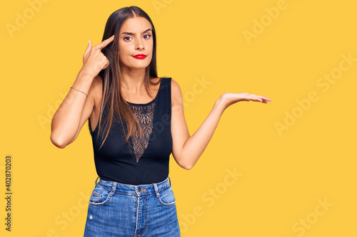 Young hispanic woman wearing casual clothes confused and annoyed with open palm showing copy space and pointing finger to forehead. think about it.