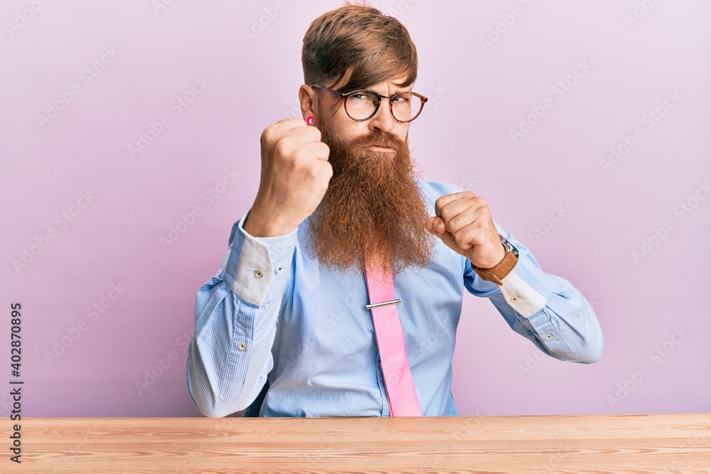 Young irish redhead man wearing business shirt and tie sitting on the table ready to fight with fist defense gesture, angry and upset face, afraid of problem Young irish redhead man wearing business shirt and tie sitting on the table ready to fight with fist defense gesture, angry and upset face, afraid of problem