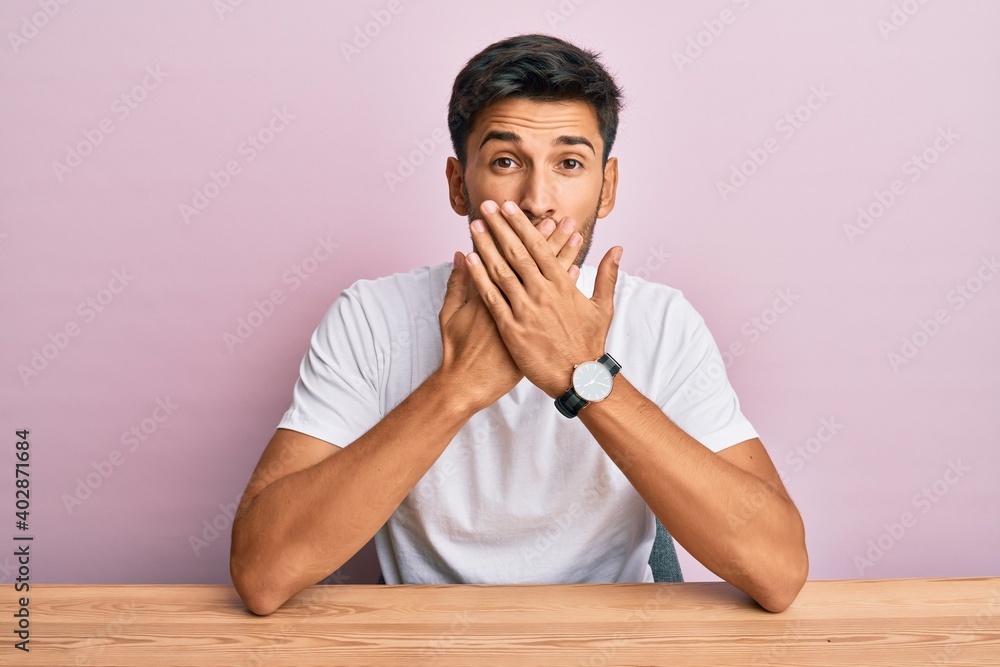 Young handsome man wearing casual white t-shirt sitting on the table shocked covering mouth with hands for mistake. secret concept. Young handsome man wearing casual white t-shirt sitting on the table shocked covering mouth with hands for mistake. secret concept.