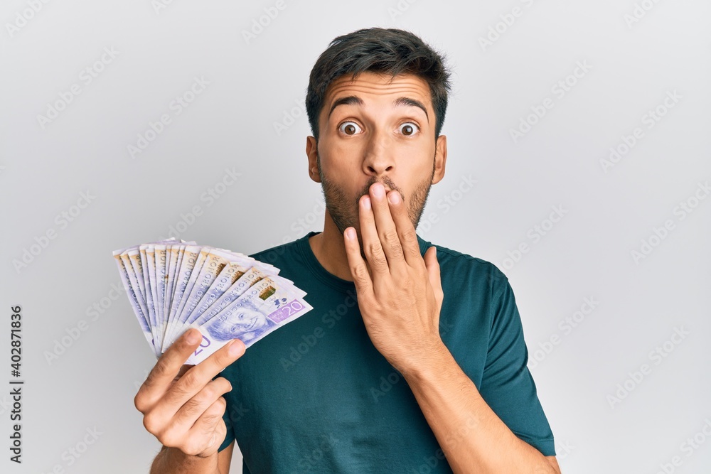 Young handsome man holding 20 swedish krona banknotes covering mouth with hand, shocked and afraid for mistake. surprised expression Young handsome man holding 20 swedish krona banknotes covering mouth with hand, shocked and afraid for mistake. surprised expression