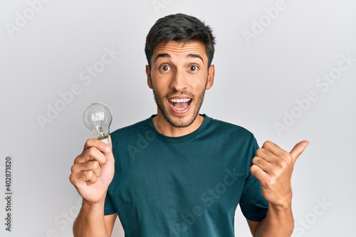 Young handsome man holding lightbulb for inspiration and idea pointing thumb up to the side smiling happy with open mouth