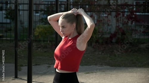 portrait of young fit blond busty woman in red top collecting hair in ponytail at sports ground. caucasian female in sportswear working out, fitness training outdoors at yard, healthy lifestyle photo