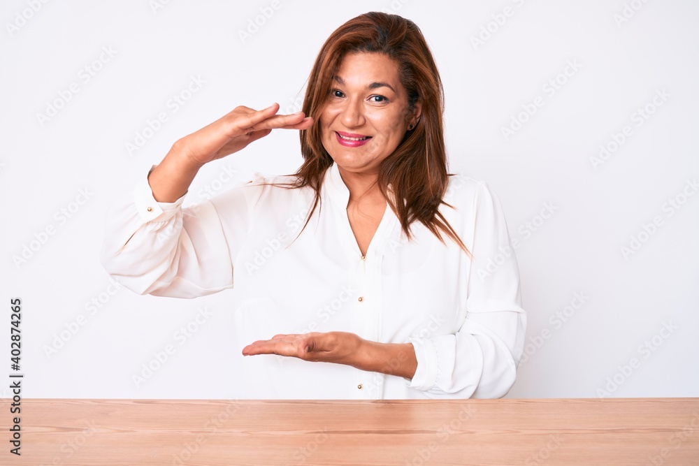 Middle age brunette hispanic business woman wearing casual white shirt sitting on the table gesturing with hands showing big and large size sign, measure symbol. smiling looking at the camera. Middle age brunette hispanic business woman wearing casual white shirt sitting on the table gesturing with hands showing big and large size sign, measure symbol. smiling looking at the camera.