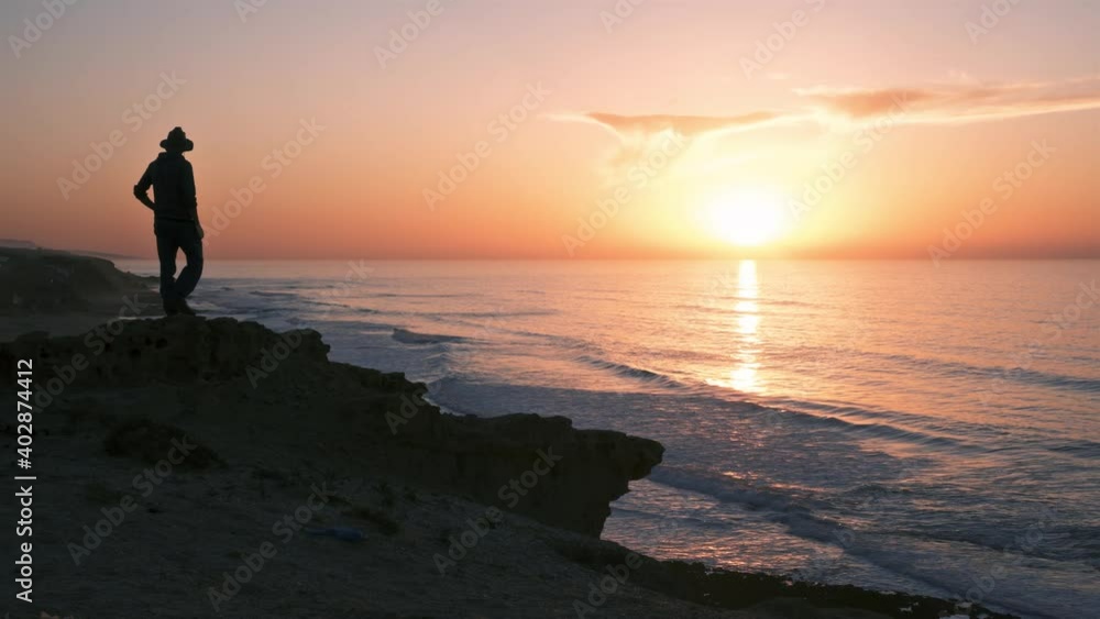 Silhouette of young man in a hat looking at beautiful sunrise in ocean coast beach in wild nature of New Zealand in peaceful summer morning Silhouette of young man in a hat looking at beautiful sunrise in ocean coast beach in wild nature of New Zealand in peaceful summer morning