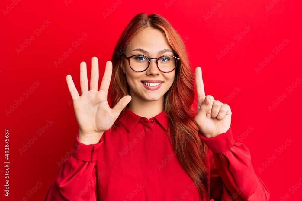 Young beautiful redhead woman wearing casual clothes and glasses over red background showing and pointing up with fingers number six while smiling confident and happy. Young beautiful redhead woman wearing casual clothes and glasses over red background showing and pointing up with fingers number six while smiling confident and happy.