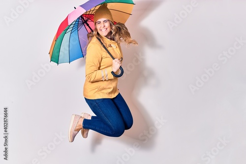Young beautiful woman wearing winter clothes smiling happy. Jumping with smile on face holding colorful umbrella over isolated white background