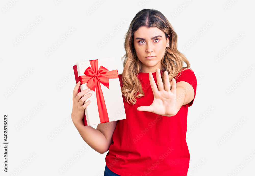 Young caucasian woman holding gift with open hand doing stop sign with serious and confident expression, defense gesture Young caucasian woman holding gift with open hand doing stop sign with serious and confident expression, defense gesture