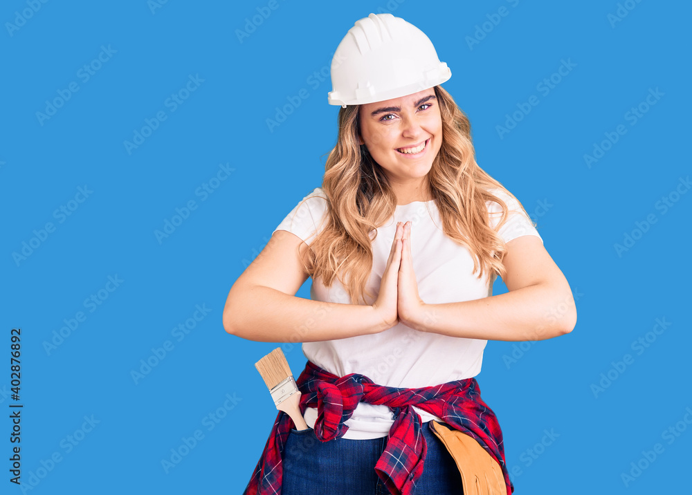 Young caucasian woman wearing security helmet praying with hands together asking for forgiveness smiling confident.