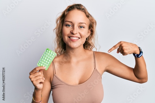 Beautiful caucasian woman holding birth control pills pointing finger to one self smiling happy and proud