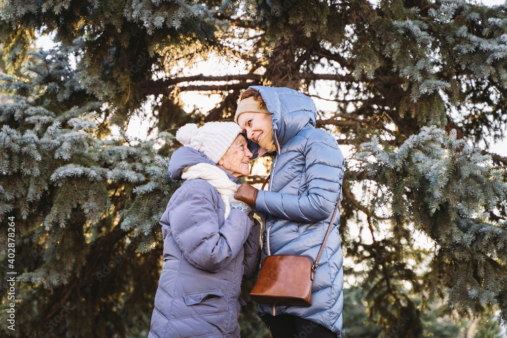 Motherhood. Theme importance visiting and spending time with old single parents during holidays. Senior mom and mature daughter happy family hug and laugh in park background Christmas tree in winter Motherhood. Theme importance visiting and spending time with old single parents during holidays. Senior mom and mature daughter happy family hug and laugh in park background Christmas tree in winter