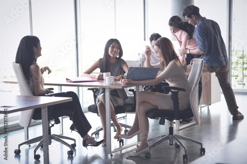 Group of young Asian business people working and communicating together while sitting at the office desk together with colleagues sitting in the background.